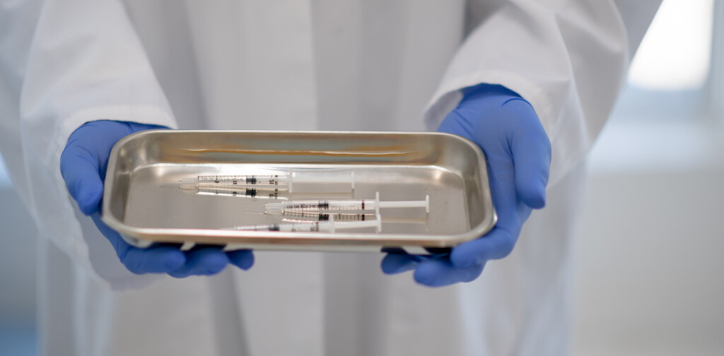 A person wearing a lab coat and blue protective gloves holds a metal tray in their hands, with three loaded syringes on it.
