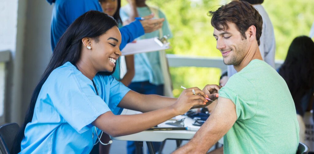 Mid adult Caucasian man smiles as African American nurse gives him a shot in his left arm. He is receiving a free medical exam and immunizations at a community outdoor free clinic. Patients and volunteers are standing in the background talking.