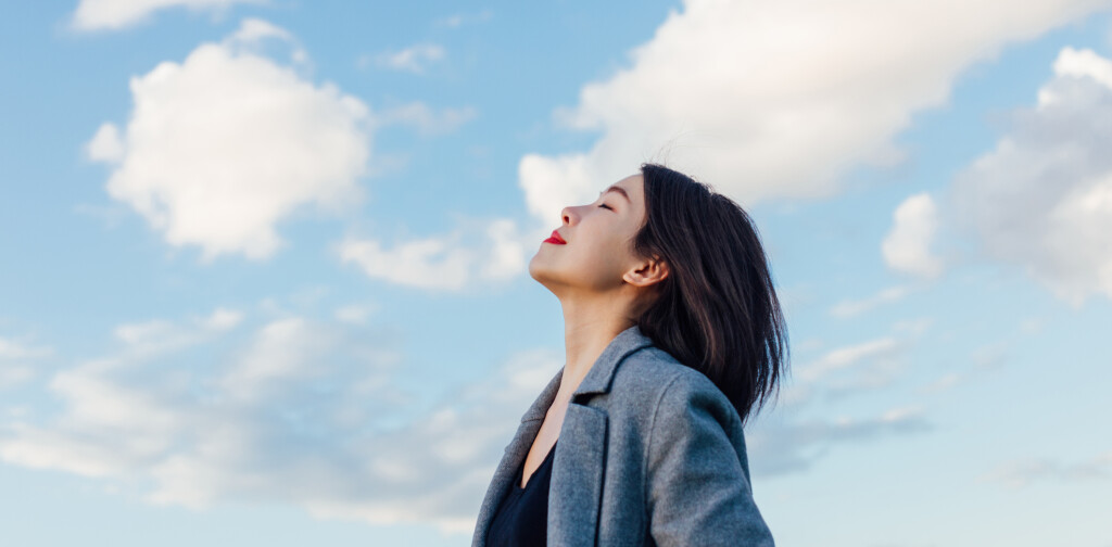 Portrait of positive young Asian woman with eye closed, enjoying sunlight under blue sky and clouds.