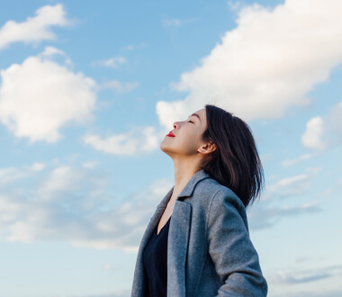 Portrait of positive young Asian woman with eye closed, enjoying sunlight under blue sky and clouds.