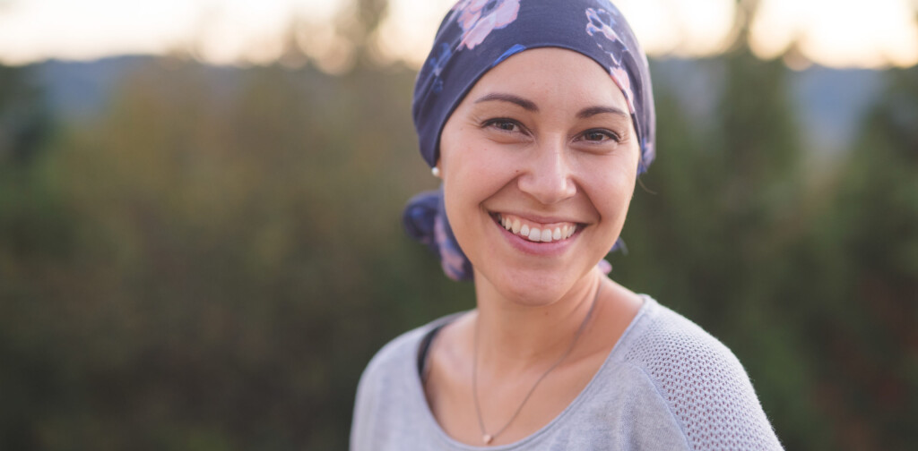 A beautiful young ethnic woman wearing a head wrap looks toward the camera and smiles radiantly. She is standing outdoors and there are mountains and trees in the background.